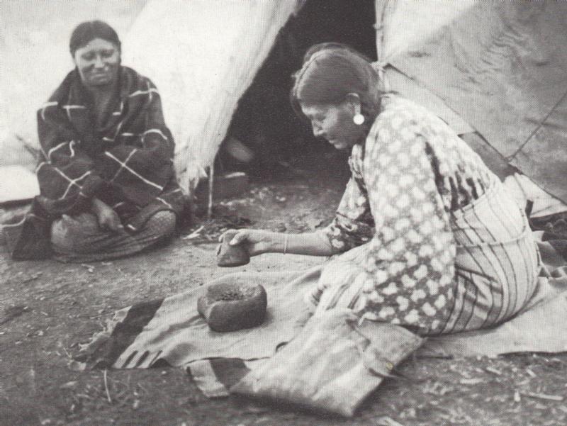 Black and white still of a woman grinding something in a bowl with a rock.