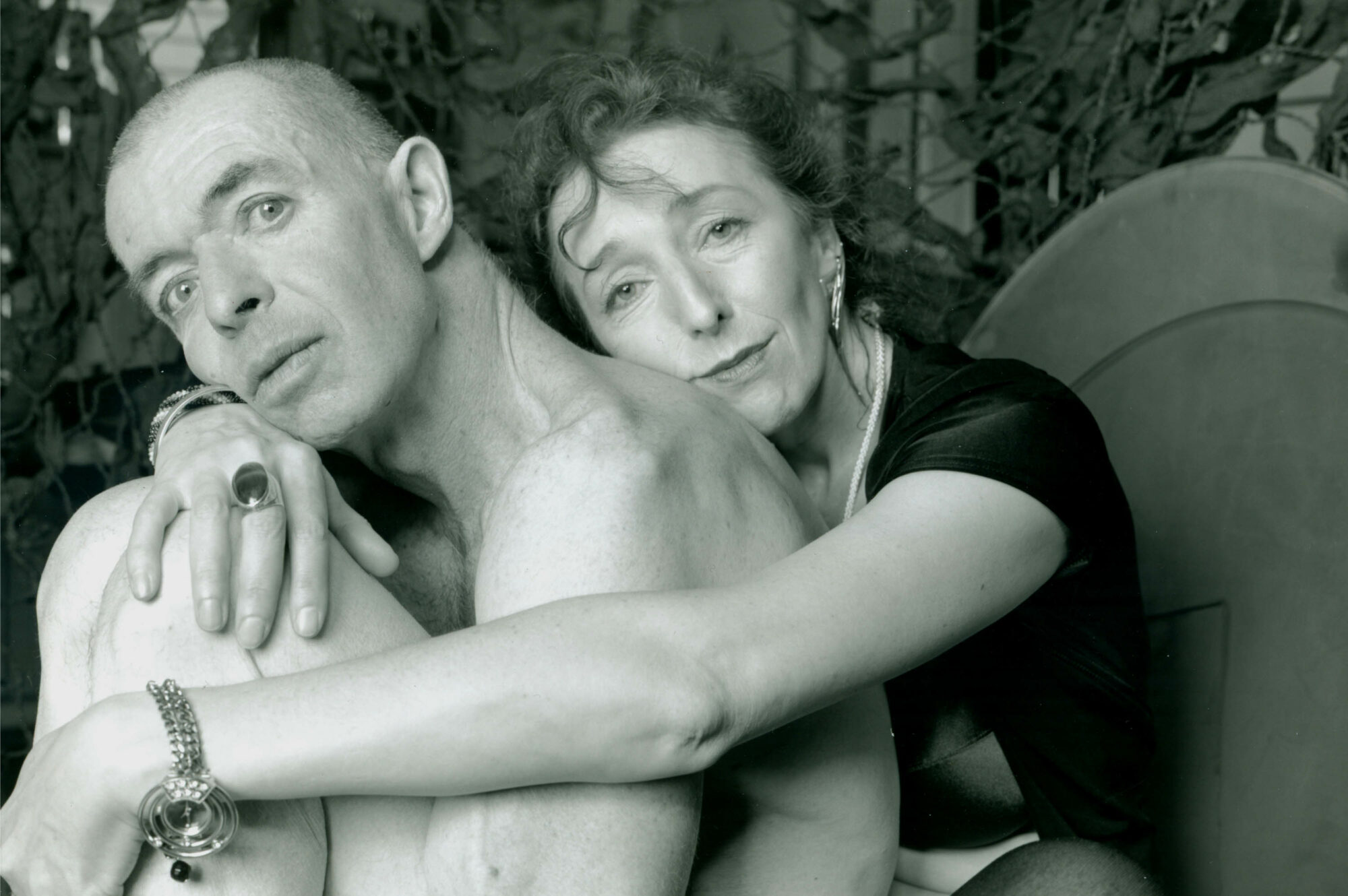 Portrait of a nude older man and a woman embracing him.