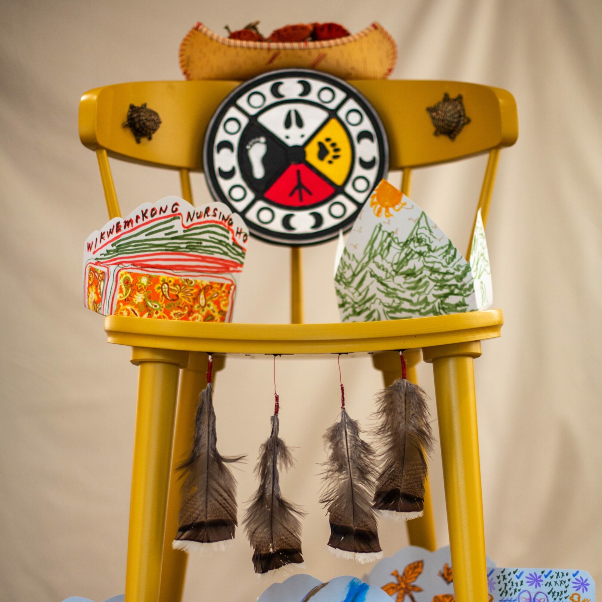 A yellow wooden chair decorated with feathers, turtles, a canoe, the Wiikwemkoong crest, and paper “Valentines.”