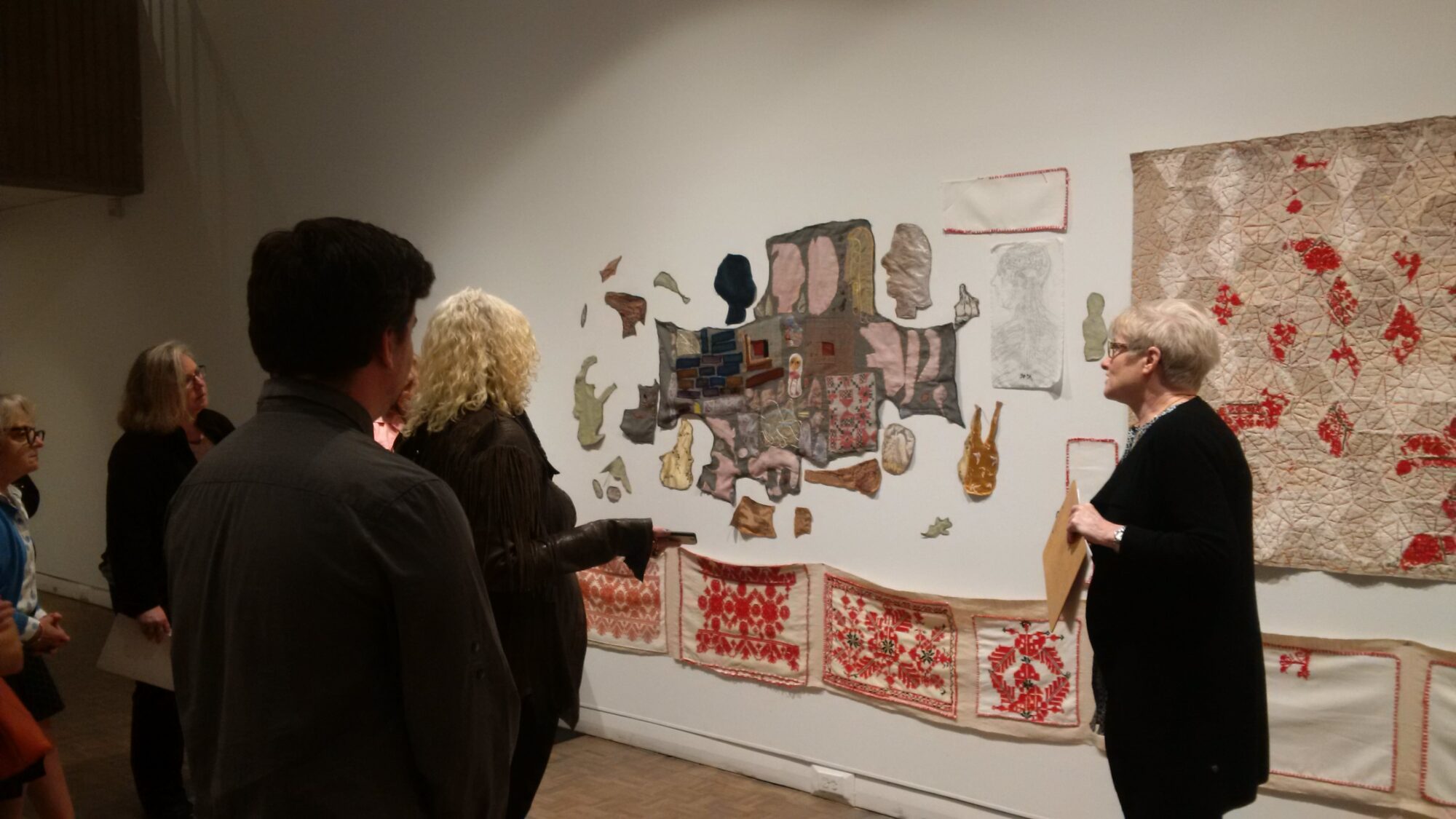 Gallery visitors looking at embroidered textiles hanging on a white wall.