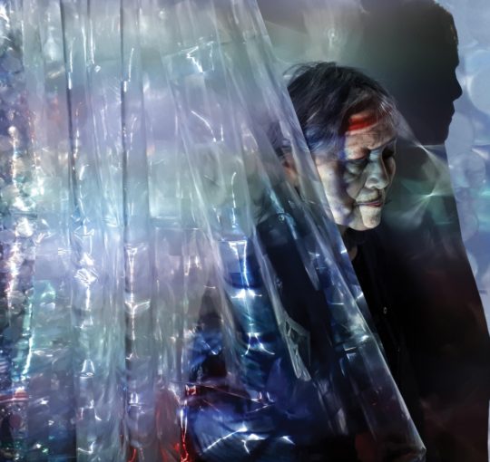 Promo image for Fault Lines of a person bathed in rainbow light from a hanging plastic sheet.
