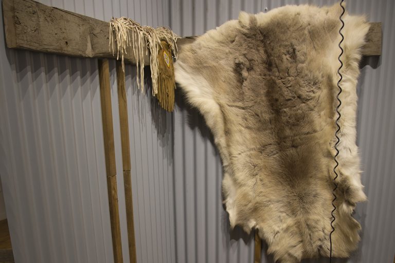 A white and grey fur pelt and a beaded pouch hang on a corrugated metal wall.