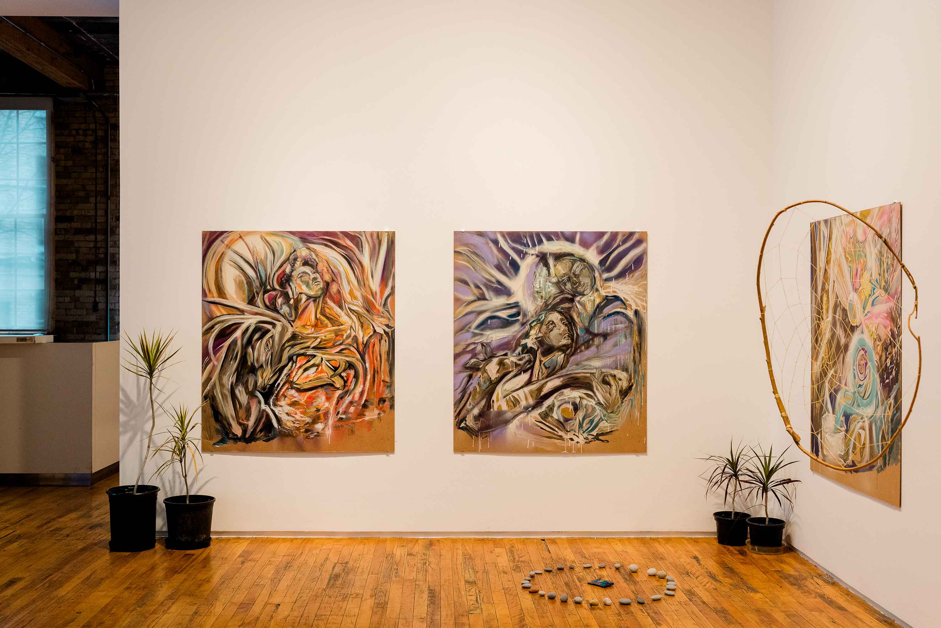 A gallery space featuring paintings, a large dream catcher, live plants, and a stone circle.
