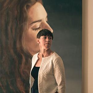 A thin white woman with a pensive face, short hair, and wearing a white blazer stands in front of a large oil painting of a woman.