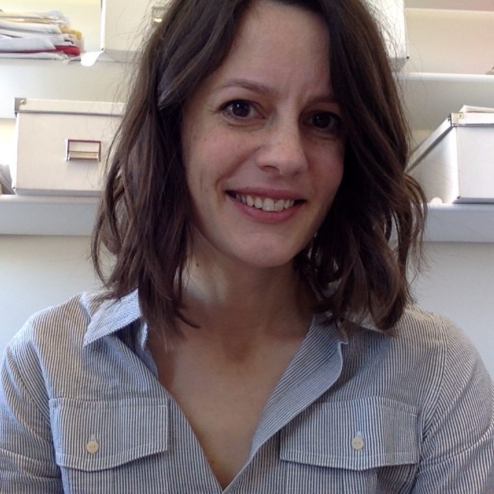 A smiling white woman with shoulder length brown hair wearing a striped grey collared shirt.